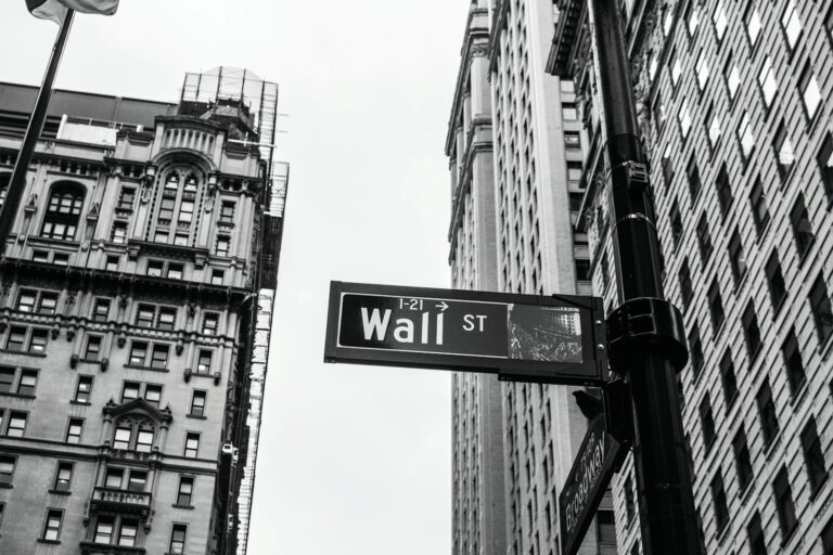 Zwart-witfoto van hoge gebouwen in een stedelijk gebied met een straatnaambord met de tekst "Wall St" prominent op de voorgrond. De lucht is bewolkt, wat een dramatisch contrast toevoegt aan de scène.