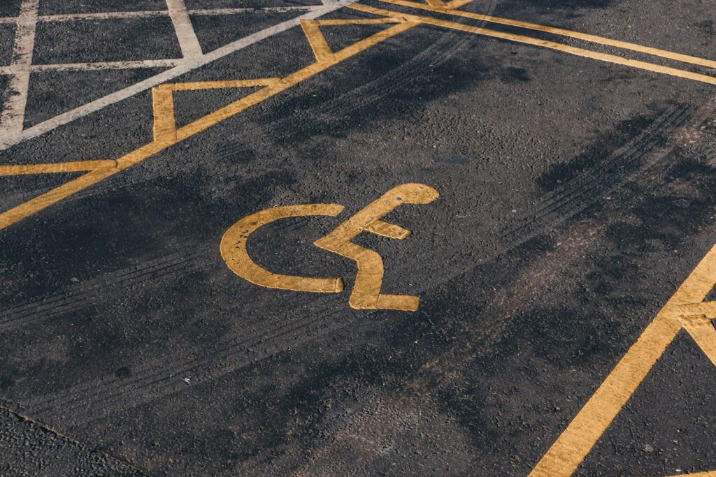 Een parkeerterrein met een geverfd geel symbool dat een aangewezen parkeerplaats voor personen met een beperking aangeeft. De grond vertoont tekenen van slijtage en vage bandensporen.