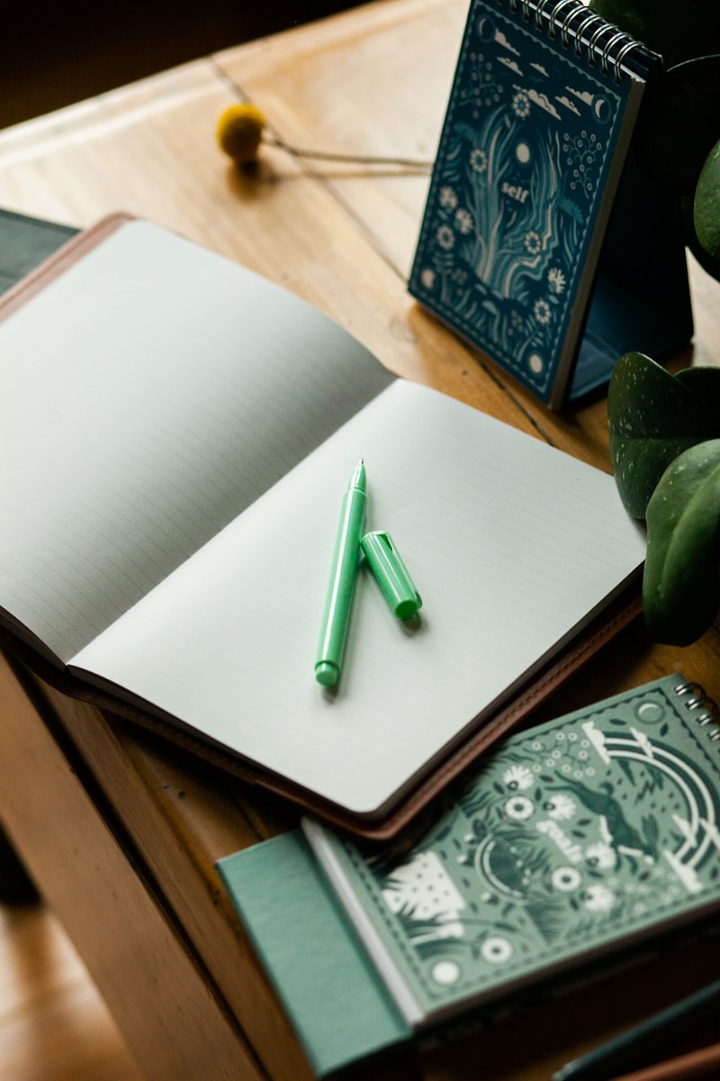 Een open notitieboekje met lege pagina's ligt op een houten tafel. Een groene pen met de dop eraf ligt op het notitieboekje. Twee spiraalgebonden notitieblokken met decoratieve omslagen liggen ernaast en een kleine plant verschijnt gedeeltelijk in het frame.