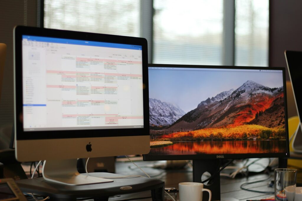 Twee computermonitoren op een bureau. Het linkerscherm toont een kalenderapplicatie en het rechterscherm toont een bureaubladachtergrond van een berglandschap. Een koffiemok staat op de voorgrond en er zijn snoeren zichtbaar rond de monitoren.