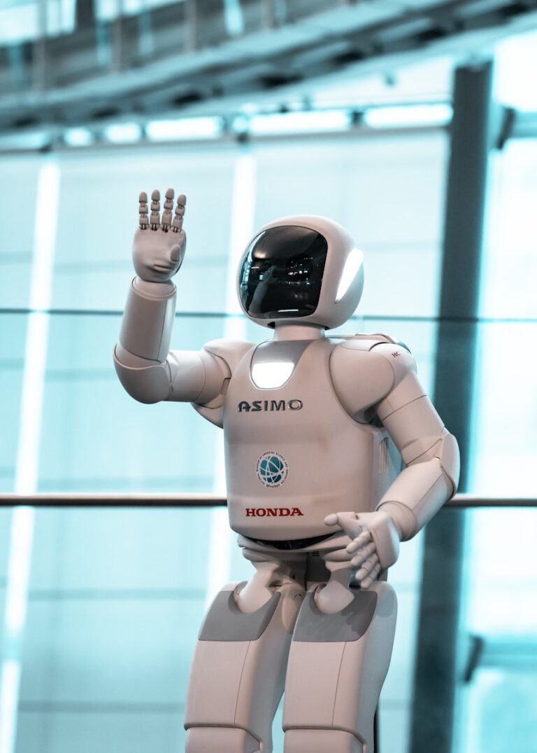 Een witte humanoïde robot met de naam ASIMO op zijn borst staat binnen op een glimmende vloer. Hij heft zijn rechterarm op, alsof hij zwaait. De achtergrond is voorzien van grote ramen waar licht doorheen filtert.