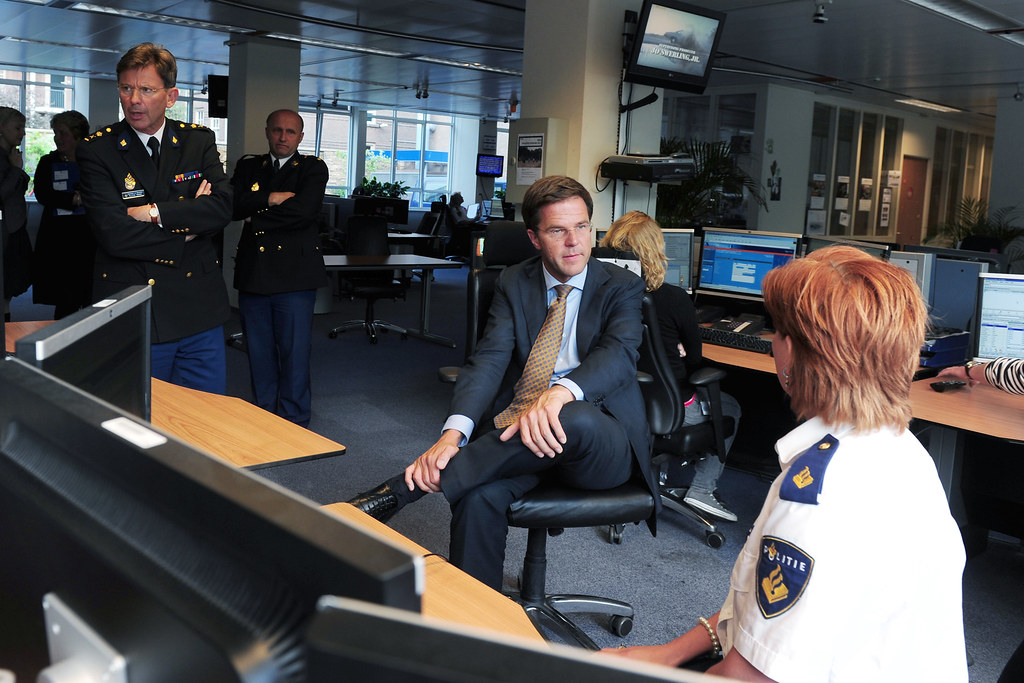 Een man in pak zit in een kantooromgeving en spreekt met een vrouw in uniform aan een bureau. Andere personen, sommigen in uniform, staan in de buurt. Computerschermen en kantoormeubilair zijn zichtbaar op de achtergrond.