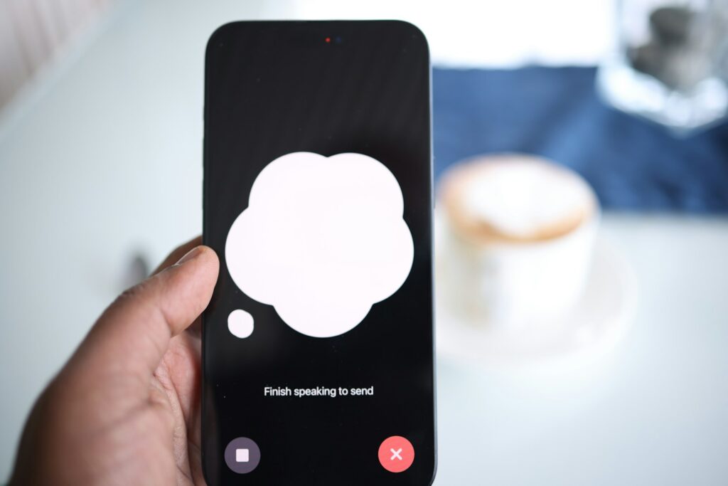 Een hand die een smartphone vasthoudt met een tekstballonpictogram op het scherm. Onder het pictogram staat de tekst "Finish speaking to send." Een wazige koffiekop is zichtbaar op de achtergrond op een tafel.