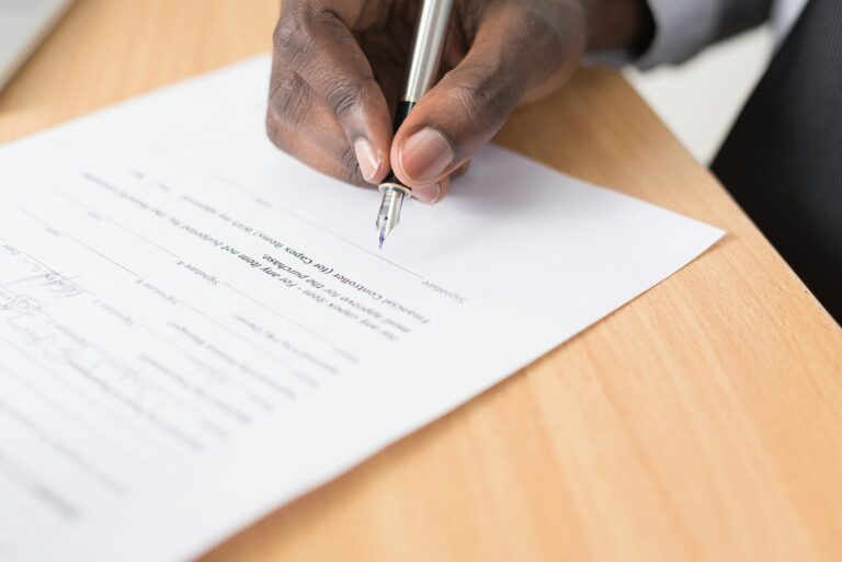 Een persoon houdt een pen vast en ondertekent een document op een houten bureau. Het document bevat gedrukte tekst en heeft onderaan een ruimte voor een handtekening. De hand van de persoon is zichtbaar, samen met een deel van een donkergekleurde mouw.