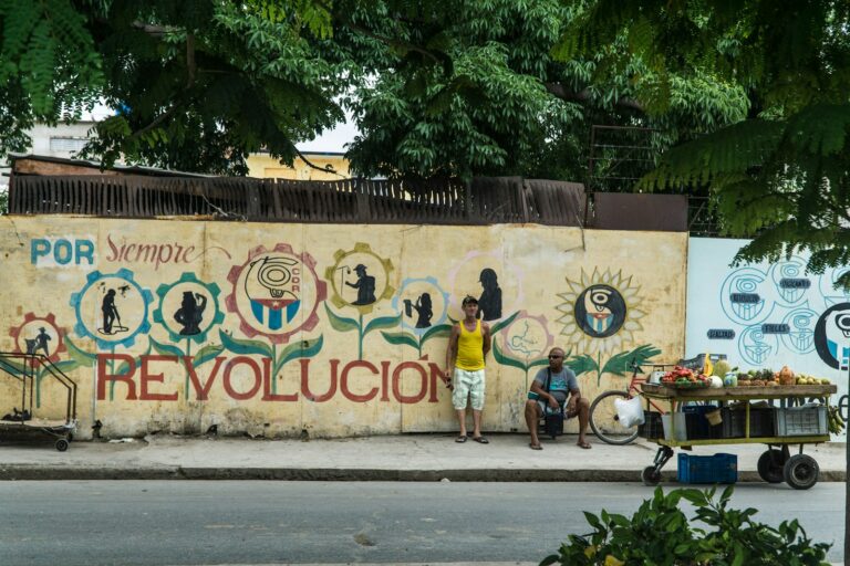 Een muurschildering met de zin "Por Siempre Revolución" toont verschillende silhouetten en abstracte ontwerpen. Vooraan staan twee mannen naast een fruitkar. Weelderige groene bomen bedekken gedeeltelijk de bovenkant van de muurschildering.