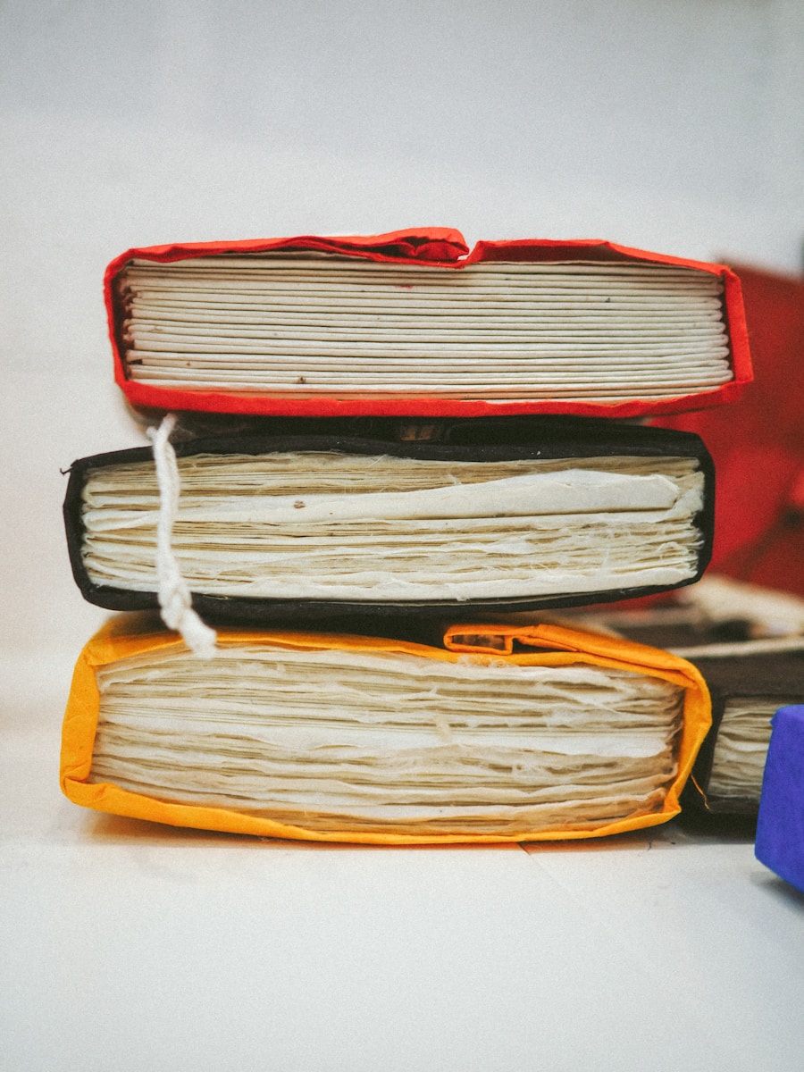 Een stapel van vier dikke, hardcover notitieboeken met getextureerde, versleten pagina's. Het bovenste boek heeft een rode cover, het tweede is zwart, het derde is oranje en een stukje van de blauwe cover van het vierde boek is zichtbaar aan de onderkant.