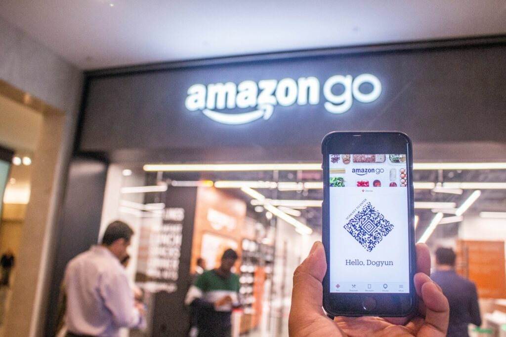 Een persoon houdt een smartphone omhoog met daarop een QR-code met de tekst "Hallo, Dogruu" voor de ingang van een Amazon Go-winkel. Er worden meerdere mensen gezien die de winkel in- en uitgaan. Het bord van de winkel is prominent zichtbaar boven de ingang.