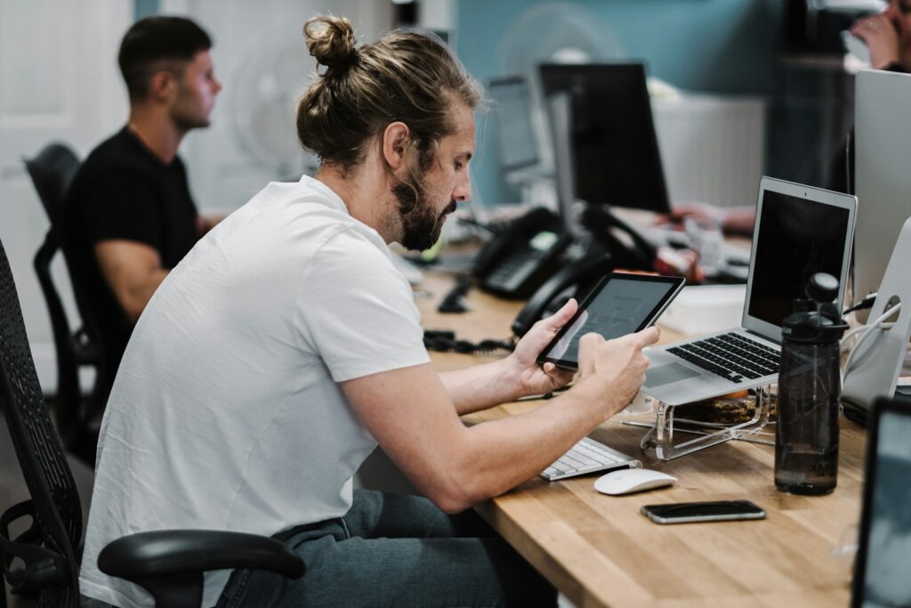 Een man met lang haar en een baard zit aan een bureau in een kantoor, met een tablet in zijn hand. Er staan meerdere computers, een waterfles en kantoorbenodigdheden op het bureau. Een andere persoon werkt op een computer op de achtergrond.