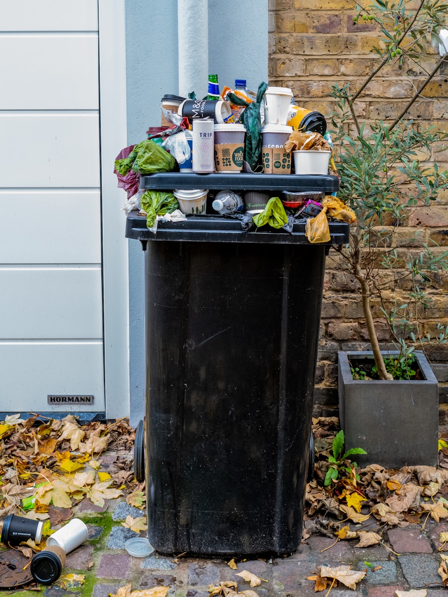 Een overvolle zwarte prullenbak is omringd door gevallen bladeren. Hij is gevuld met verschillende items, waaronder karton, blikjes en plastic zakken, en staat bij een bakstenen muur en een kleine potboom.