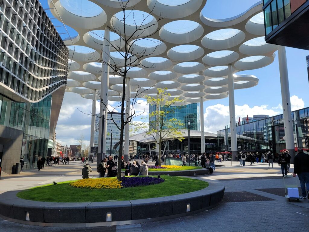 Een modern stedelijk plein bij station Utrecht met een unieke overheadstructuur met cirkelvormige uitsparingen. Mensen verzamelen zich rond aangelegde groene ruimtes met gele en paarse bloemen. Omringende glazen gebouwen weerspiegelen de lucht, wat bijdraagt aan de hedendaagse sfeer.