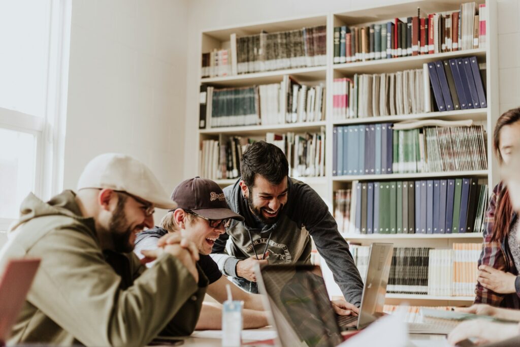 Een groep van vier mensen zit aan een tafel in een bibliotheek of kantoor, kijkt naar een laptop en lacht. Boekenplanken vol met boeken en mappen staan op de achtergrond. De sfeer lijkt ongedwongen en vriendelijk.