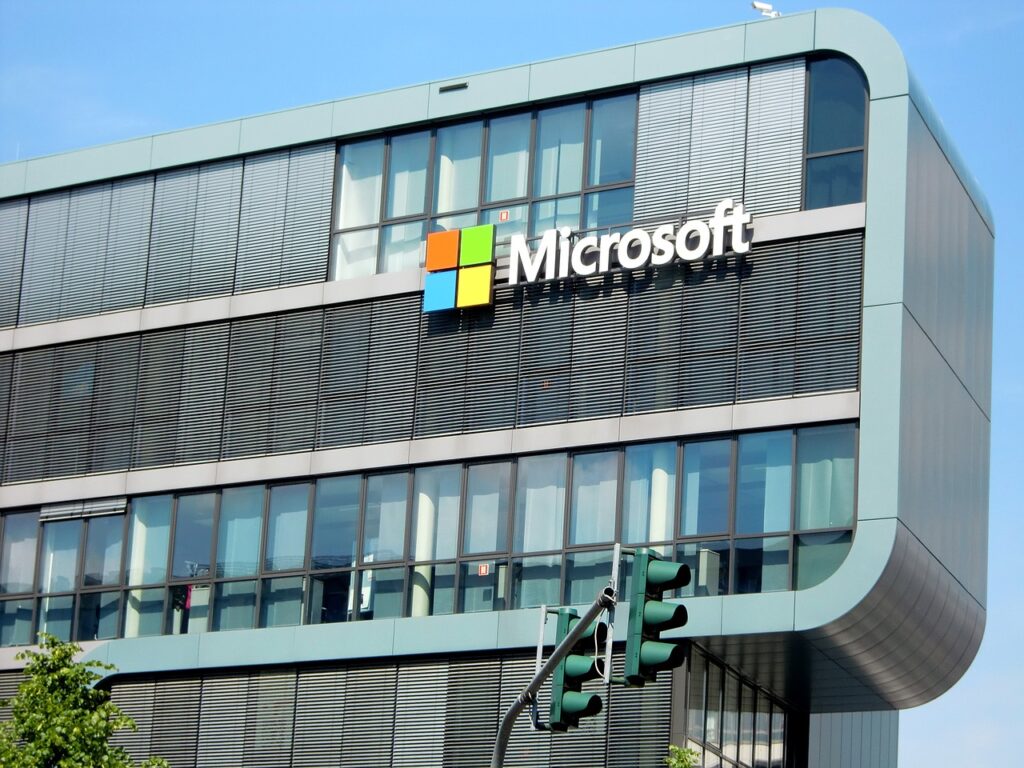 Buitenaanzicht van een modern kantoorgebouw met het Microsoft-logo op de gevel. Het gebouw heeft grote ramen en een gebogen ontwerp. De lucht is helder en blauw.