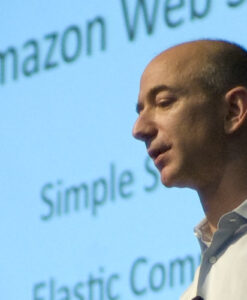 Een persoon (Jeff Bezosa) spreekt voor een projectiescherm met tekst waarop onder andere "Amazon Web Services", "Simple S" en "Elastic Com" staan. De persoon staat en profil afgebeeld, met een overhemd met kraag.