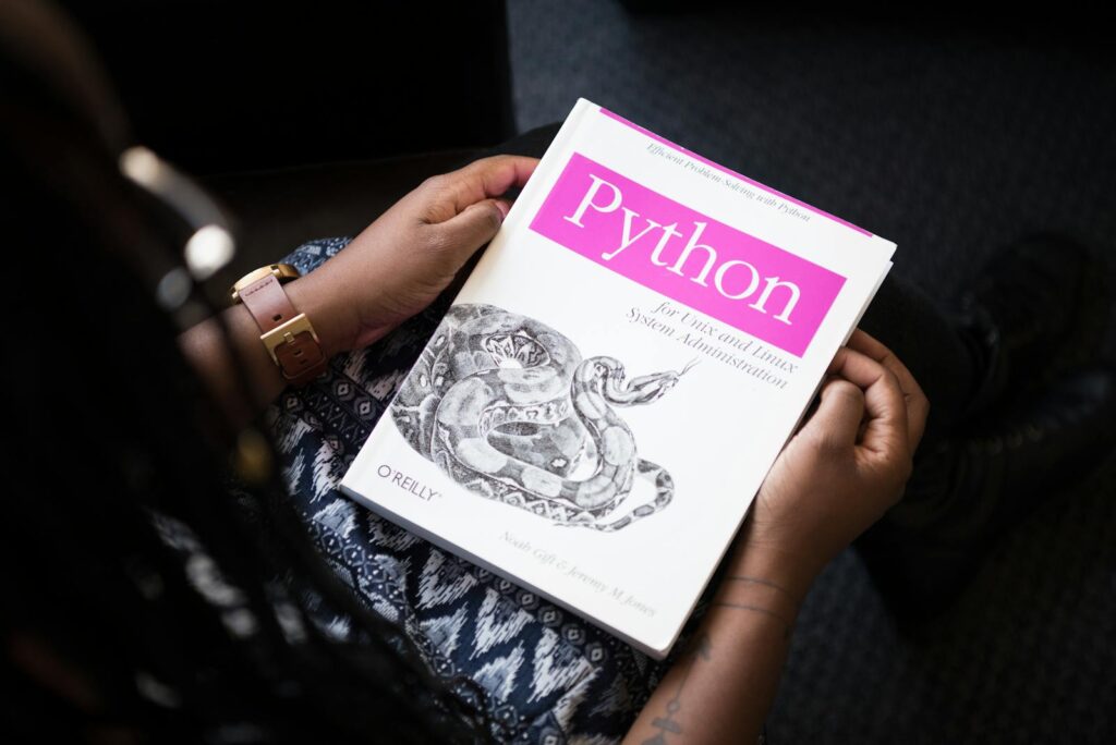 Een persoon houdt een boek vast met de titel "Python for Unix and Linux System Administration" van O'Reilly. De cover toont een tekening van een slang. De persoon draagt een patroonoutfit en een horloge.