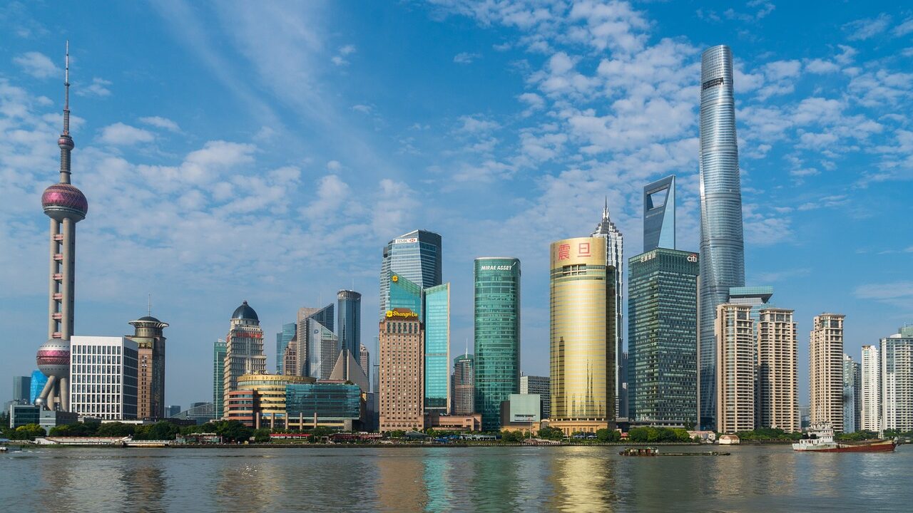 Een skyline van Shanghai met hoge, moderne wolkenkrabbers langs een waterkant, onder een blauwe lucht met wolken. Opvallende bouwwerken zijn onder andere een opvallende toren met een rond observatiedek. Het water op de voorgrond weerspiegelt de gebouwen.