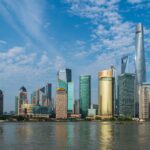Een skyline van Shanghai met hoge, moderne wolkenkrabbers langs een waterkant, onder een blauwe lucht met wolken. Opvallende bouwwerken zijn onder andere een opvallende toren met een rond observatiedek. Het water op de voorgrond weerspiegelt de gebouwen.