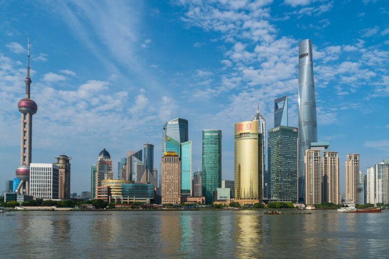 Een skyline van Shanghai met hoge, moderne wolkenkrabbers langs een waterkant, onder een blauwe lucht met wolken. Opvallende bouwwerken zijn onder andere een opvallende toren met een rond observatiedek. Het water op de voorgrond weerspiegelt de gebouwen.