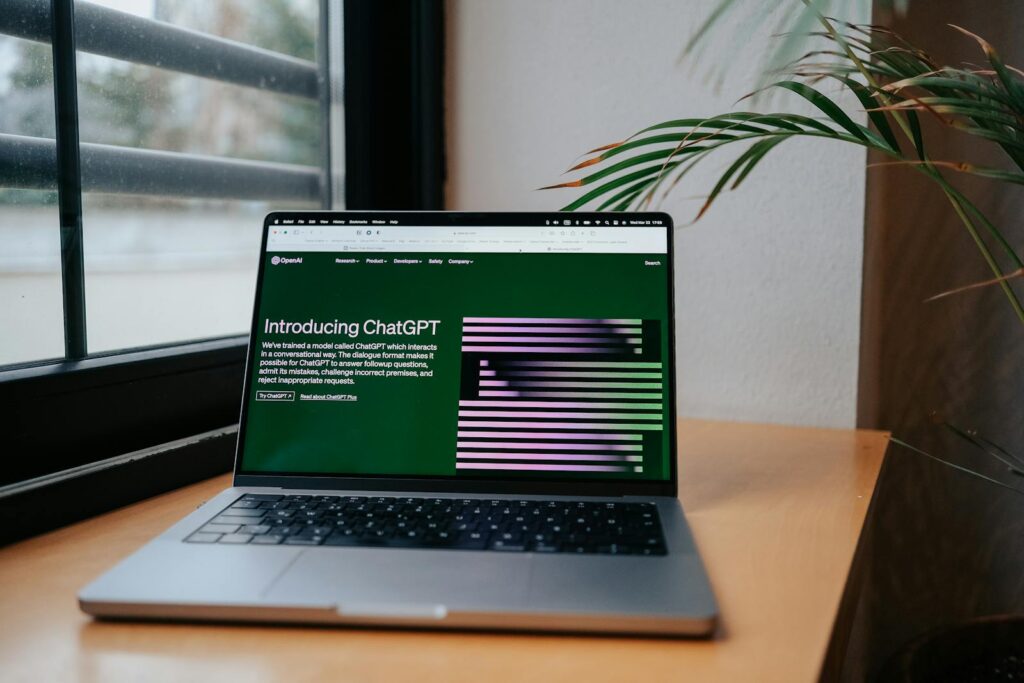 Een laptop staat op een houten bureau bij een raam met tralies. Het scherm toont een webpagina met de titel "Introducing ChatGPT", met groene en paarse afbeeldingen. Een plant met lange bladeren is gedeeltelijk zichtbaar aan de rechterkant van de laptop.