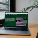 Een laptop staat op een houten bureau bij een raam met tralies. Het scherm toont een webpagina met de titel "Introducing ChatGPT", met groene en paarse afbeeldingen. Een plant met lange bladeren is gedeeltelijk zichtbaar aan de rechterkant van de laptop.