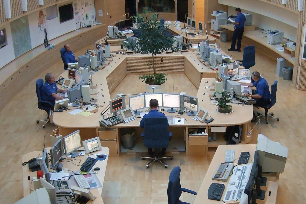 De afbeelding toont een luchtfoto van een controlekamer met meerdere werkplekken die in een U-vorm zijn opgesteld. Meerdere mensen zitten aan bureaus met computerschermen en er staat een grote plant in het midden van de kamer.