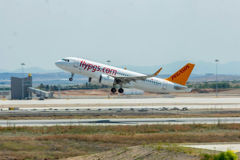 Een vliegtuig van Pegasus Airlines stijgt op vanaf een landingsbaan op het vliegveld. Het vliegtuig, met de website "flypgs.com" op de zijkant, stijgt op in de lucht. Op de achtergrond zijn luchthavengebouwen en een heldere lucht te zien.