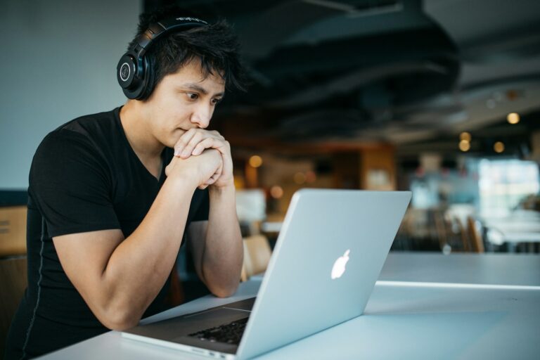 Een persoon met een koptelefoon zit aan een tafel en kijkt aandachtig naar een laptopscherm. De setting lijkt een informele binnenruimte met wazige achtergrondelementen.