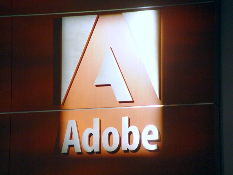 De afbeelding toont een muur met een verlicht Adobe-logo. Het logo bestaat uit een grote letter "A" boven het woord "Adobe" in witte 3D-letters op een bruine achtergrond. Licht schijnt op het bovenste gedeelte van het logo.