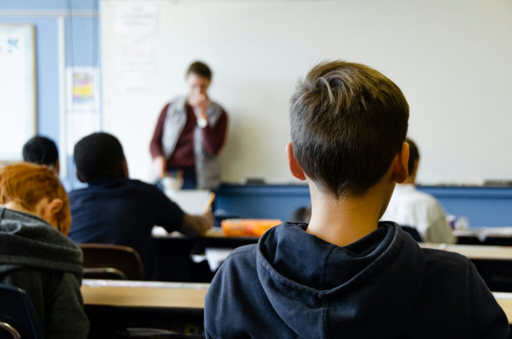 Een klaslokaalscène met studenten die aan een bureau zitten en een leraar voor een whiteboard aankijken. De focus ligt op een student met kort haar, die een donkere hoodie draagt. De leraar houdt een stift in de hand en lijkt te instrueren.