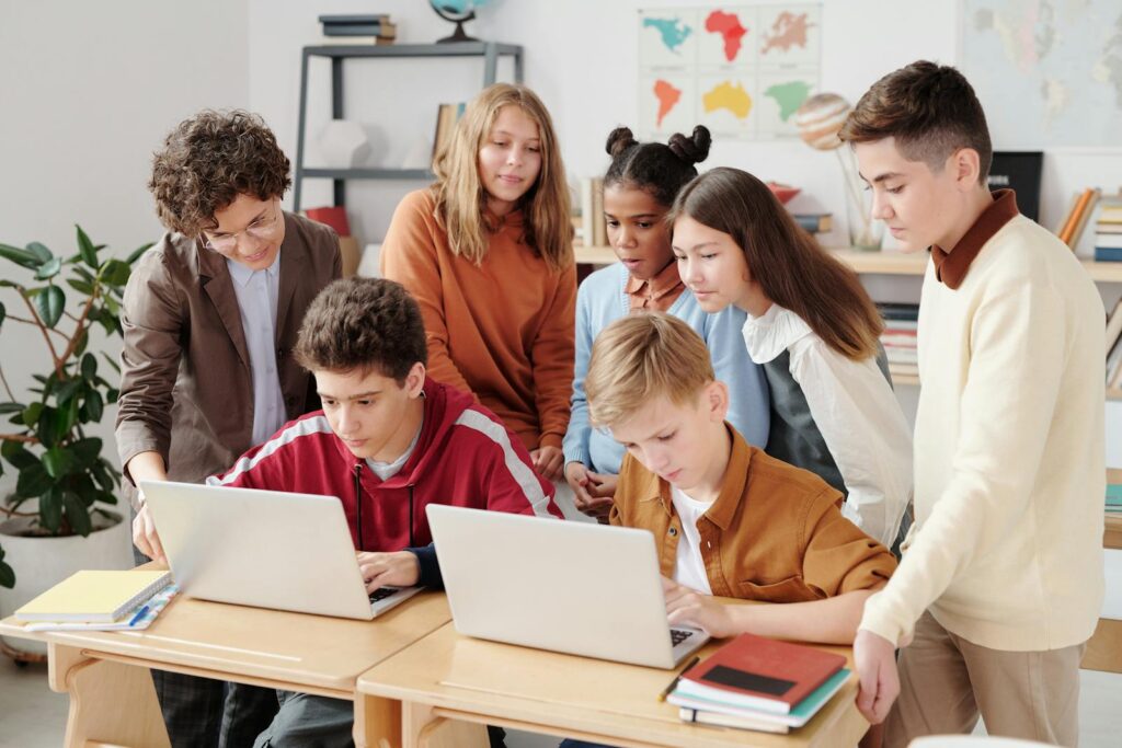 Een groep studenten verzameld rond twee bureaus, elk met een laptop. Een volwassene helpt de studenten terwijl anderen nauwlettend toekijken. Op de achtergrond zijn kaarten en boekenplanken te zien.