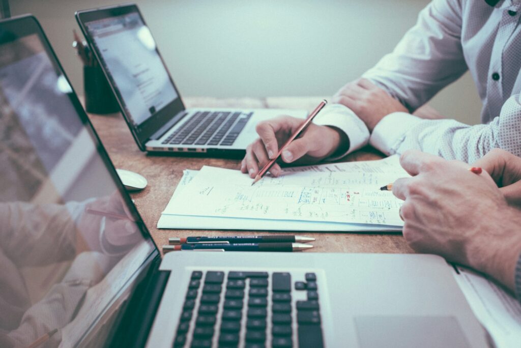 Twee mensen zitten aan een tafel met laptops en papieren. Eén persoon houdt een rood potlood vast en wijst naar een document met schetsen en aantekeningen. Drie andere potloden rusten op de tafel.