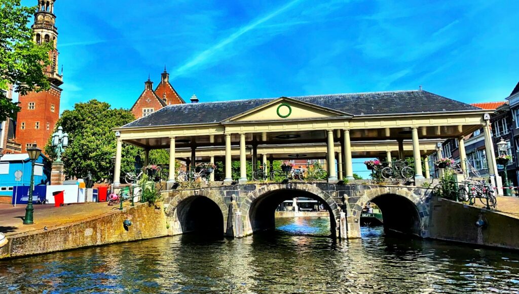 Een historische stenen brug met drie bogen ondersteunt een overdekte voetgangersroute versierd met bloemen en fietsen. Rode bakstenen gebouwen en een klokkentoren zijn zichtbaar op de achtergrond onder een helderblauwe lucht. Leiden.