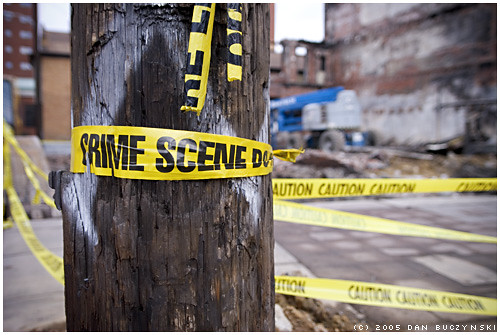 Een houten paal is omwikkeld met geel crime scene tape voor een gedeeltelijk gesloopt gebouw. Extra waarschuwingstape is zichtbaar op de achtergrond en er is bouwapparatuur aanwezig.