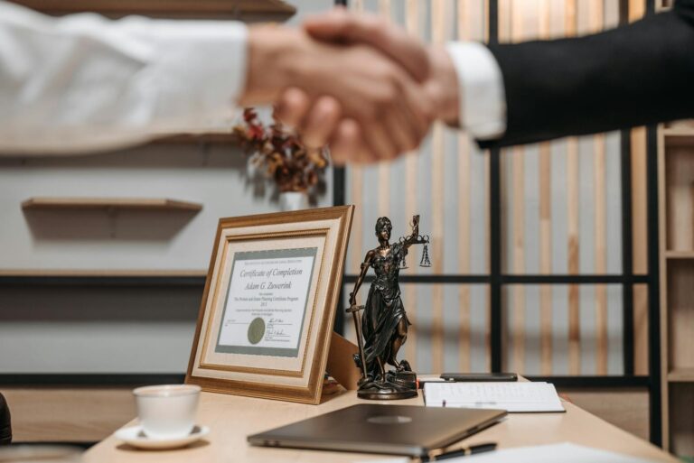 Twee mensen schudden elkaar de hand boven een bureau met daarop een ingelijst certificaat, een klein beeldje van Vrouwe Justitia, een laptop, een document en een kop koffie. Op de achtergrond staan planken met decoratie-items.