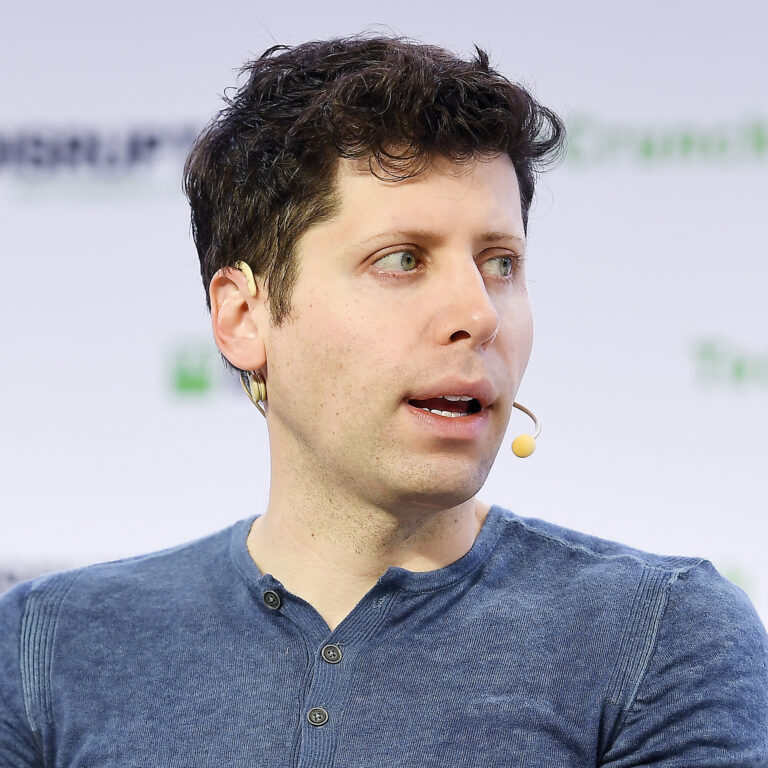 Sam Altman, CEO van OpenAi, die een blauw shirt en een microfoonheadset draagt, spreekt. Ze zitten tegen een achtergrond met onduidelijke, herhaalde tekst.