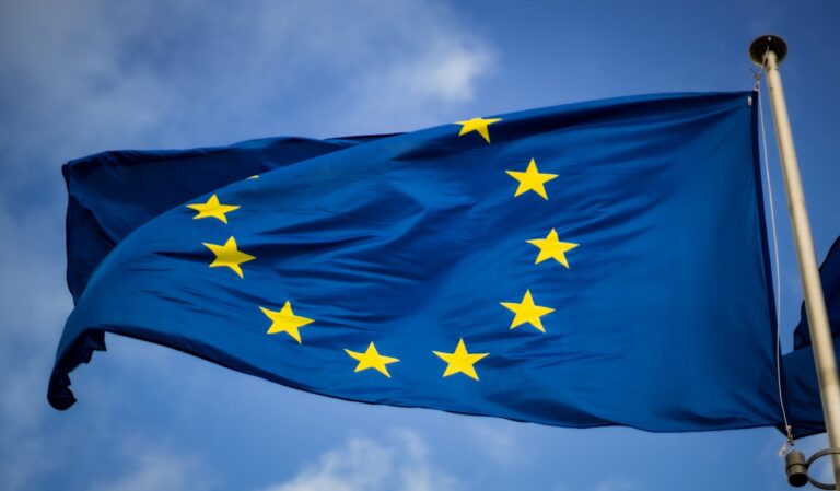 De afbeelding toont de vlag van de Europese Unie, met een cirkel van twaalf gouden sterren op een blauwe achtergrond. De vlag wappert tegen een gedeeltelijk bewolkte lucht.