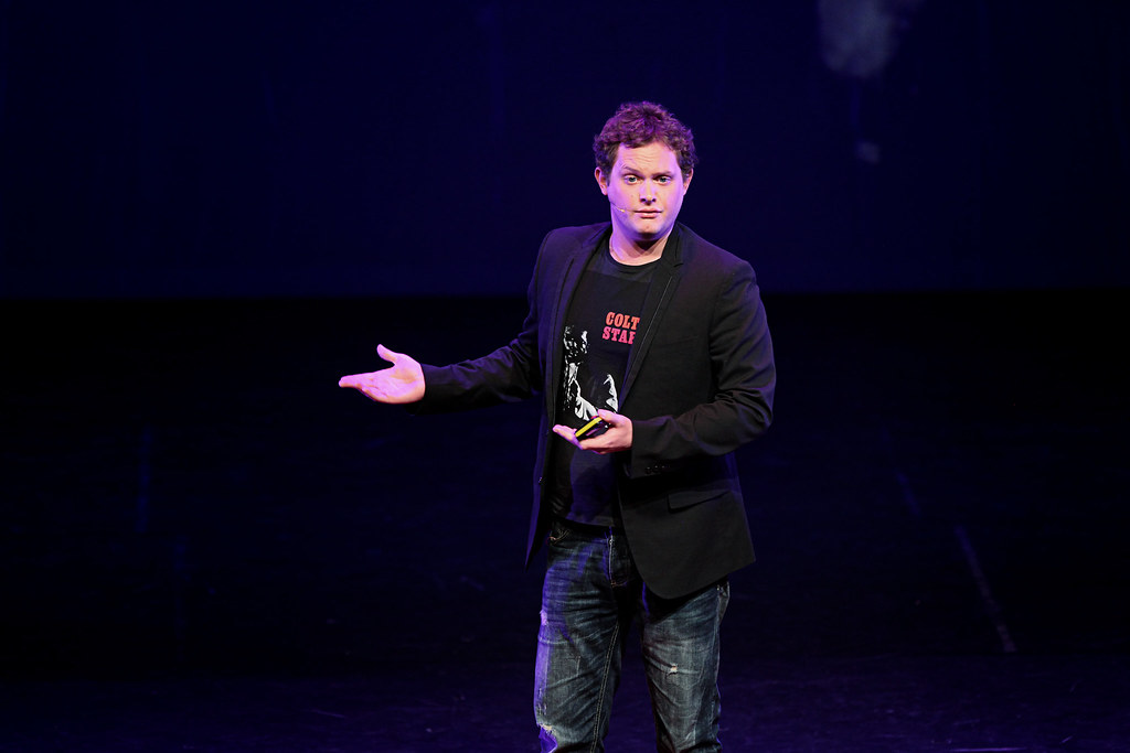 Een persoon (Diederik Jekel) staat op het podium met een donkere achtergrond, draagt een zwarte blazer, een grafisch T-shirt en jeans. Ze hebben één hand open in een gebarende pose.