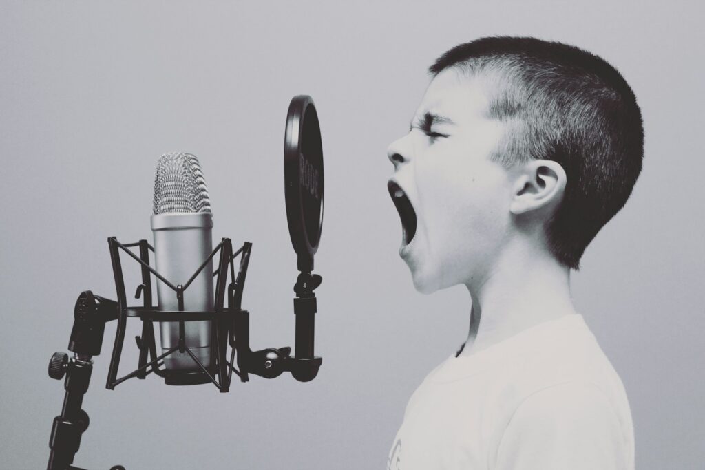 Een zwart-witafbeelding van een jonge jongen die hartstochtelijk zingt of schreeuwt in een microfoon. De jongen heeft kort haar en draagt een lichtgekleurd shirt. De microfoon is op een standaard gemonteerd met een popfilter ervoor.