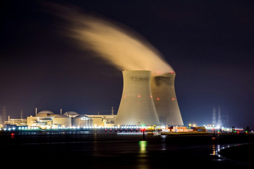 Een nachtelijke scène van een kerncentrale met twee grote koeltorens die stoom uitstoten. De faciliteit wordt verlicht door kunstlicht en is omgeven door elektrische infrastructuur, met een donkere lucht op de achtergrond.