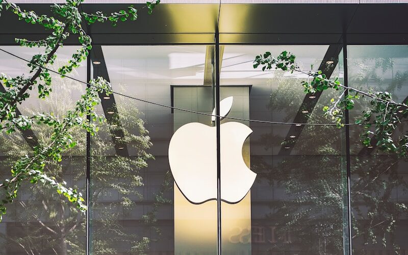 De afbeelding toont een grote glazen winkelpui met een prominent wit Apple-logo in het midden. Groene boomtakken omlijsten het uitzicht, weerspiegeld op het glas, wat een natuurlijk element toevoegt aan de stedelijke omgeving. Het interieur van de winkel is zichtbaar door het glas, met een strak, modern ontwerp.