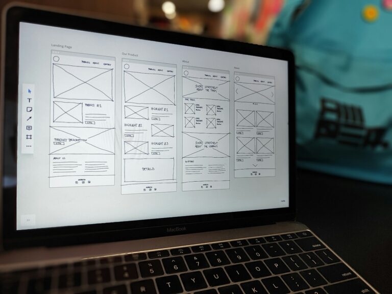 Een laptop met een digitaal wireframe-ontwerp met meerdere webpagina-indelingen. Het scherm toont secties met de labels "Landing Page", "Our Product" en "About". Er is een wazige blauwe achtergrond zichtbaar.