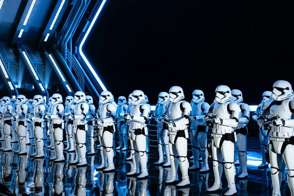 Een groep stormtroopers in witte harnassen staat in formatie in een futuristische, slecht verlichte kamer met blauwe en zwarte accenten. Ze houden blasters vast en lijken in rijen opgesteld te staan. De scène doet denken aan een sciencefictionsetting.