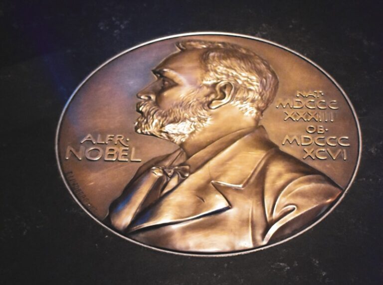 Een bronzen medaille met een reliëf van een bebaarde man in pak, met het opschrift "Alfr. Nobel". De medaille is voorzien van Romeinse cijfers, data en details op de achtergrond.