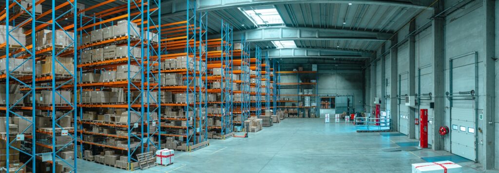 Een groot magazijninterieur met hoge blauwe en oranje planken vol met dozen en pallets. De betonnen vloer is grotendeels leeg, wat leidt naar hoge ramen die natuurlijk licht binnenlaten. Brandveiligheidsapparatuur is zichtbaar op de muren.