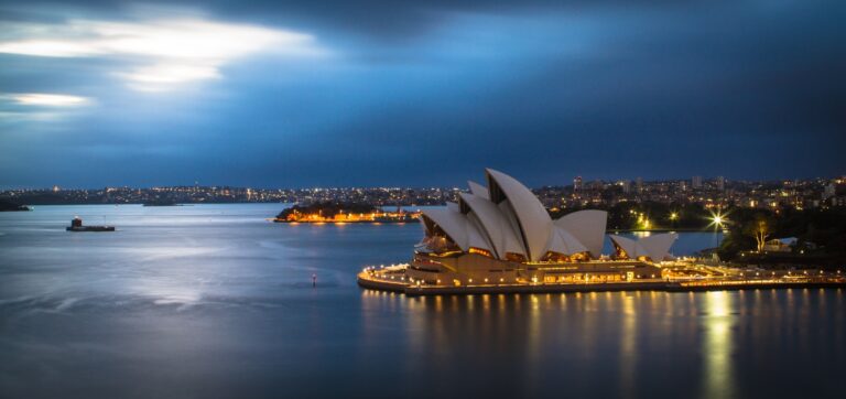 De afbeelding toont het Sydney Opera House verlicht in de nacht, omringd door water. Stadslichten zijn zichtbaar op de achtergrond onder een bewolkte lucht.