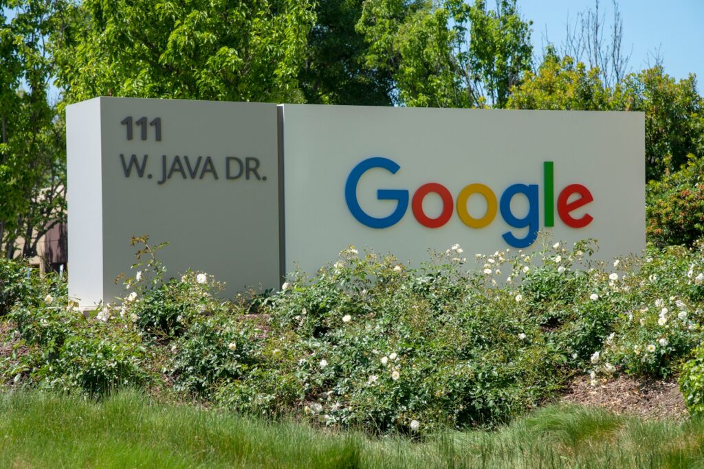 Bord met "111 W. Java Dr." en het Google-logo in kleurrijke letters, omgeven door groen en witte bloemen.