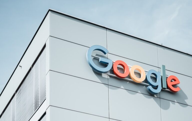 De afbeelding toont een hoek van een modern gebouw met het Google-logo in kleurrijke letters bovenaan. De gevel bestaat uit strakke, lichtgrijze panelen met smalle verticale ramen. De lucht erboven is helder en gedeeltelijk zichtbaar, wat bijdraagt aan de strakke, minimalistische look.