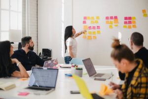 Een groep mensen zit rond een tafel met laptops en notitieboekjes, terwijl een vrouw naar een whiteboard wijst dat bedekt is met plaknotities. De kamer is licht met grote ramen die natuurlijk licht binnenlaten.