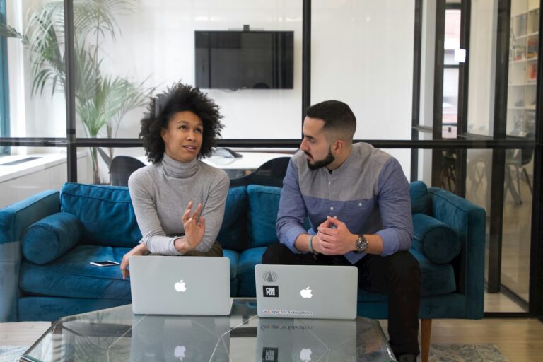 Twee mensen zitten op een blauwe bank met laptops voor zich, in gesprek. De kamer heeft een glazen wand, een tv en een plant op de achtergrond. Een smartphone ligt naast de laptop aan de linkerkant.