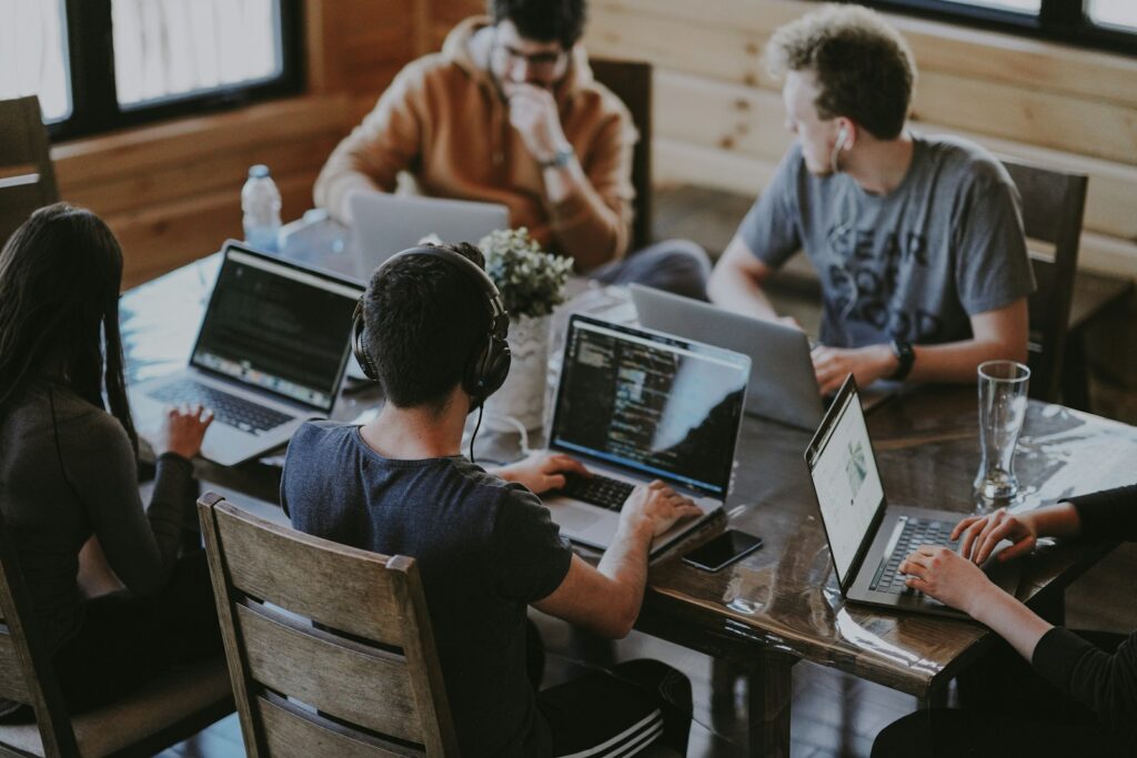 Vijf mensen zitten rond een tafel en werken op laptops. De groep lijkt geconcentreerd, sommige personen dragen koptelefoons. De omgeving suggereert een collaboratieve of co-workingruimte met een houten interieur en natuurlijk licht dat door een raam naar binnen komt.