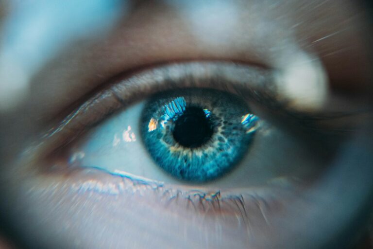 Close-up afbeelding van een blauw oog met gedetailleerde irispatronen. Lichtreflecties zijn zichtbaar op het hoornvlies, wat helderheid toevoegt. De omliggende huid is enigszins onscherp, wat de details van het oog benadrukt.